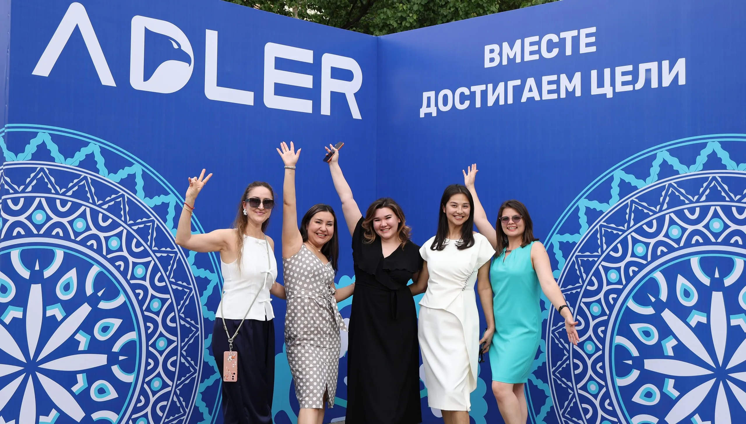 Пять сотрудниц Adler с поднятыми руками у фирменной фотостены с надписью «Вместе достигаем цели»